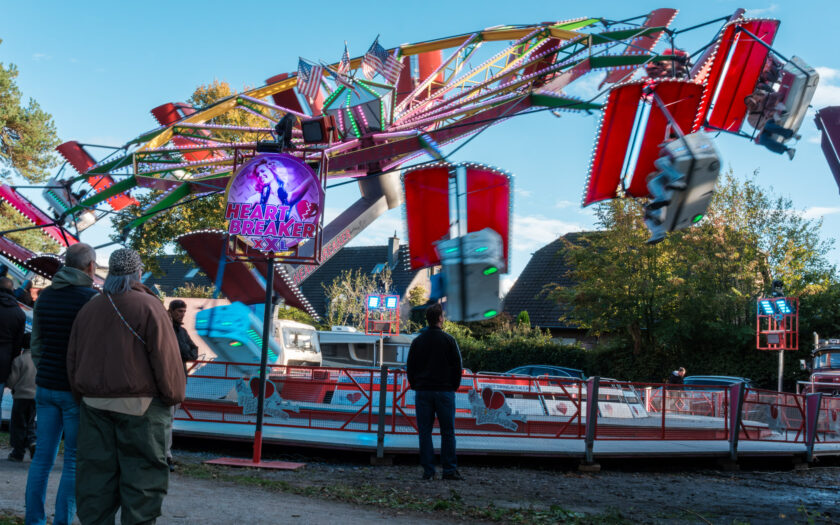Besucher auf der Hünxer Honigkirmes 2026 zwischen Ständen und Fahrgeschäft. Die Personen sind von hinten oder unscharf zu sehen.